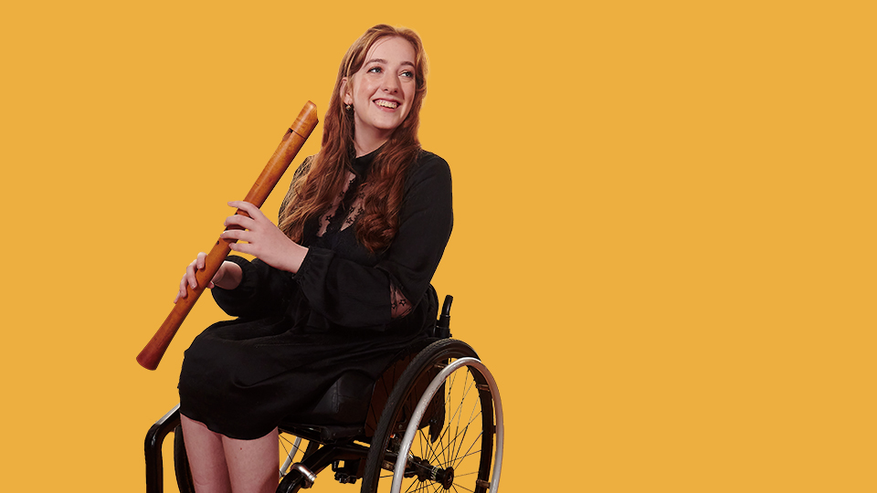 A women wearing a black dress, smiling,  holding a historical clarinet, sitting in a wheelchair.